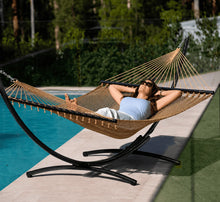 Load image into Gallery viewer, Person relaxing in a tan Caribbean rope hammock on a black steel arc stand beside a swimming pool.