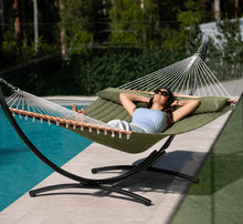 Load image into Gallery viewer, Person relaxing in an olive king quilted hammock with matching pillow on a black steel arc stand beside a swimming pool.