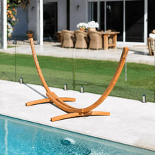 Load image into Gallery viewer, Angled view of the stained timber arc hammock stand and wide support feet beside a swimming pool