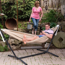 Load image into Gallery viewer, Man relaxing in a Sand hammock on a black steel stand, with a woman standing nearby in a garden.