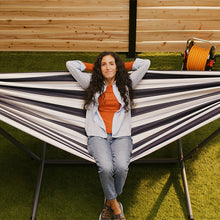 Load image into Gallery viewer, Woman sitting in a striped hammock on grass with wooden wall in the background