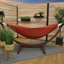 Load image into Gallery viewer, Crimson Sunbrella® hammock on a solid pine arc stand in a timber-screened space with potted plants and a patterned rug.