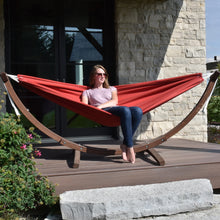 Load image into Gallery viewer, Person relaxing in a Crimson hammock on a solid pine arc stand on an outdoor deck beside a stone wall.