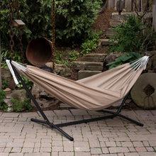 Load image into Gallery viewer, Sand double hammock on a black steel stand on a paved garden patio beside stone steps.
