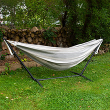 Load image into Gallery viewer, White hammock on a black stand in a garden setting with trees and logs in the background.
