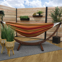 Load image into Gallery viewer, Sunset striped hammock on a solid pine arc stand in a timber-screened space with potted plants and a patterned rug.