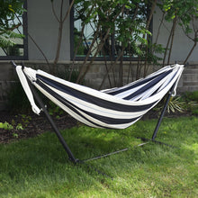 Load image into Gallery viewer, A striped hammock with black and white bands is suspended on a black metal stand in a backyard setting with green grass, trees, and a building in the background.