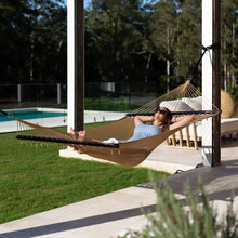 Load image into Gallery viewer, Person reclining in a tan Caribbean rope hammock suspended beside a covered patio and garden