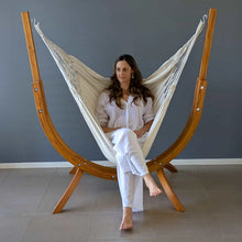 Load image into Gallery viewer, Person seated in a cream hammock chair suspended from the Coastal Lounger timber stand indoors