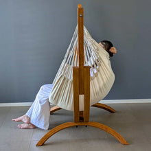 Load image into Gallery viewer, Side view of a person reclining in the cream hanging chair on the Coastal Lounger timber stand