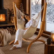 Load image into Gallery viewer, Person relaxing in a cream hanging chair on the Coastal Lounger timber stand beside a fireplace