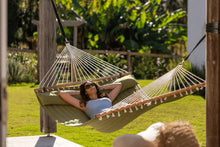 Load image into Gallery viewer, Person reclining on the matching pillow in an olive quilted hammock beside a sunny garden