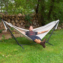 Load image into Gallery viewer, A man relaxes in a beige fabric hammock suspended on a black metal stand in a grassy backyard with a log wall and trees behind him.