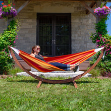 Load image into Gallery viewer, Front view of a person relaxing in a Sunset hammock on a pine arc stand outside a stone house.