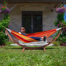 Load image into Gallery viewer, Person reclining in a Sunset striped hammock on a solid pine arc stand on a lawn beside flowering plants.
