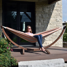 Load image into Gallery viewer, Person relaxing in a Cameo hammock on a solid pine arc stand on an outdoor deck beside a stone wall.