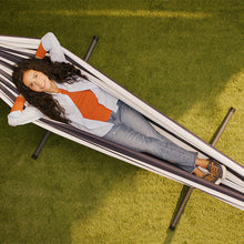 Load image into Gallery viewer, A woman relaxes in a striped white and black outdoor hammock on a green grass lawn. 