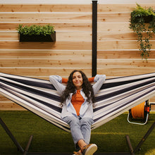 Load image into Gallery viewer, A woman relaxes in a striped hammock suspended between a wooden fence and a black metal frame, with artificial grass below and potted plants on the wall.