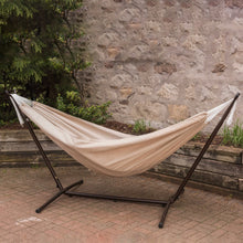Load image into Gallery viewer, Sand Sunbrella® hammock on a black steel stand on a brick patio beside a stone wall.